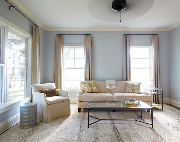 A traditional living room in beige with blue walls and each window has a tall set of beige drapes
