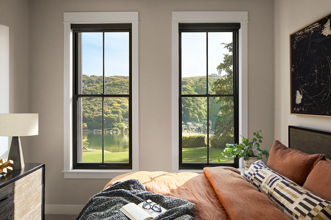 A modern bedroom featuring two black-framed windows with interior window trim, showcasing a view of greenery.
