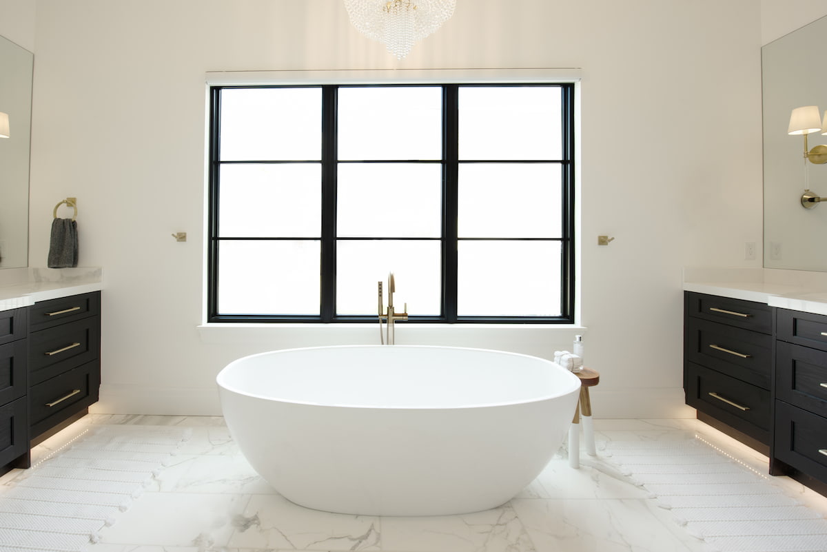 A black picture window sits over a white freestanding bathtub, with black vanities facing each other on opposite sides of the bathroom.