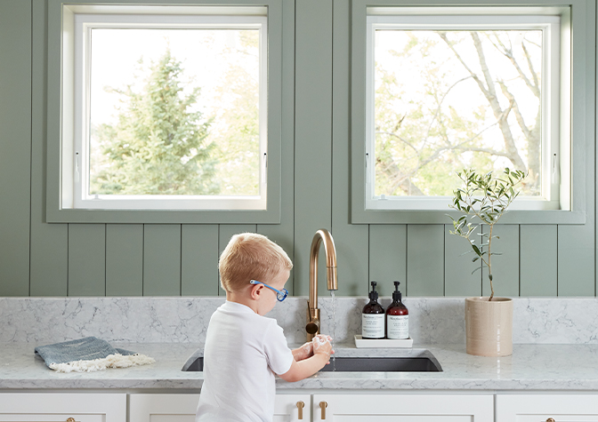 small child washing his hands at a sink underneath two awning windows.