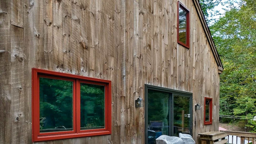 Exterior of a wooden house featuring red windows surrounded by greenery.