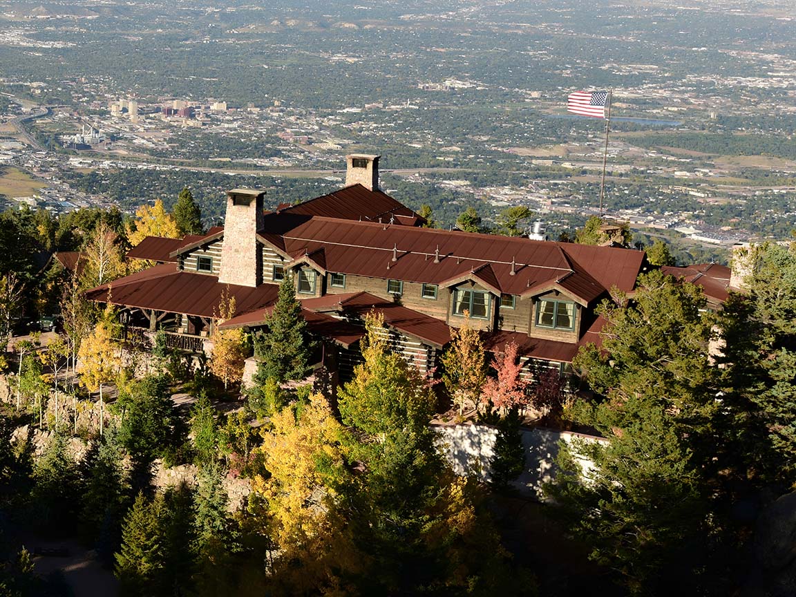 Large mountain cabin with multiple windows and a red roof surrounded by autumn trees and overlooking a city.