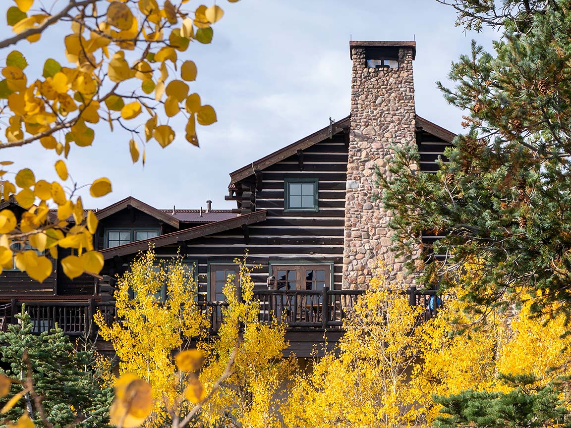 A rustic log cabin with multiple windows and a stone chimney, surrounded by autumn trees with yellow leaves.