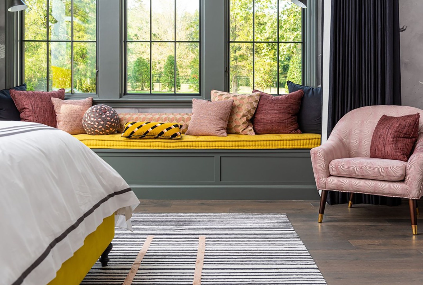 A modern bedroom with pink and yellow décor, and three windows above a seating area.