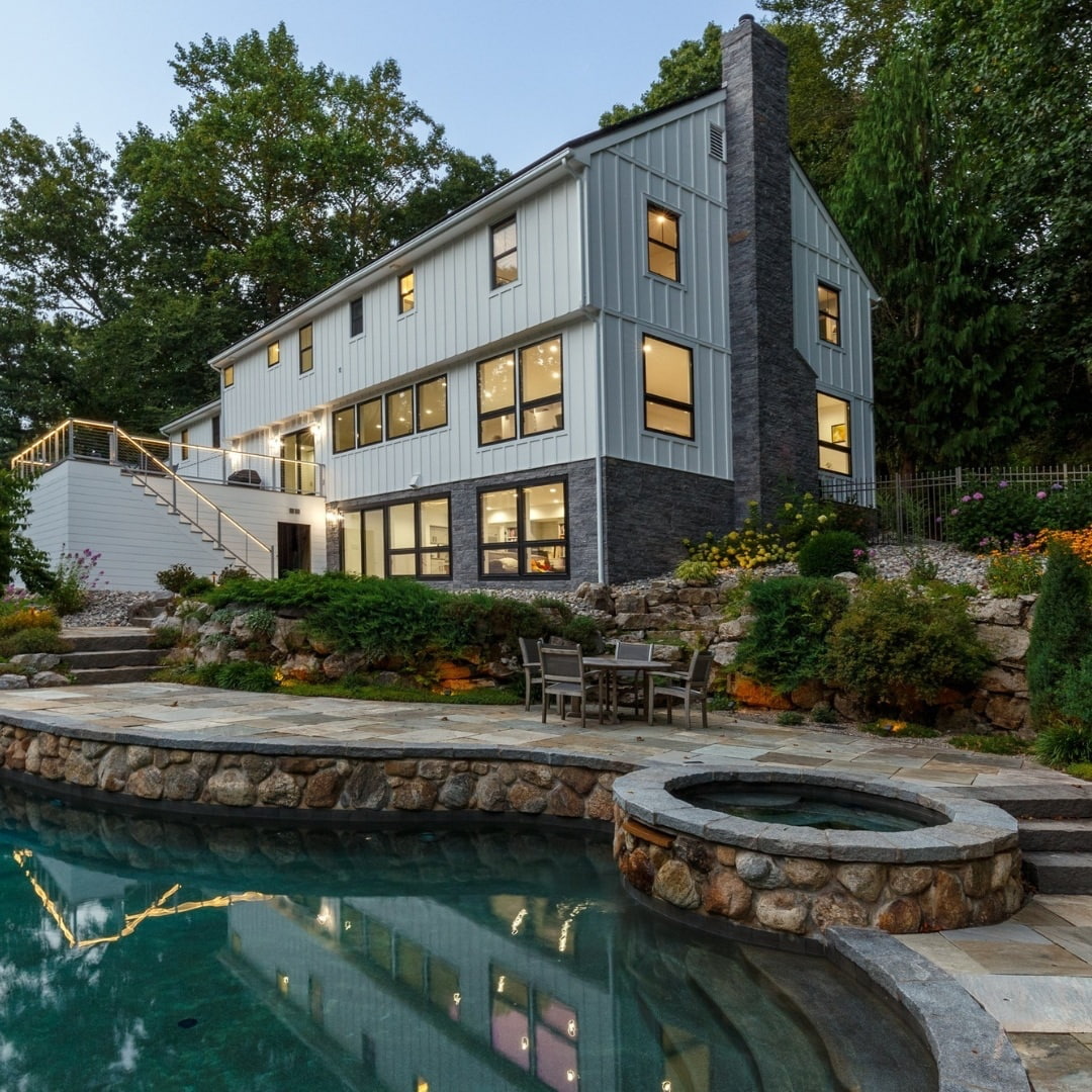 The backyard of a farmhouse-style home has basement windows and a door that overlook the pool, hot tub and patio area. 