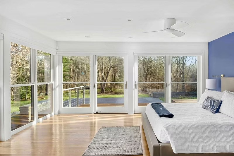 A large bedroom has walls of white windows and sliding patio doors that overlook an outdoor deck.
