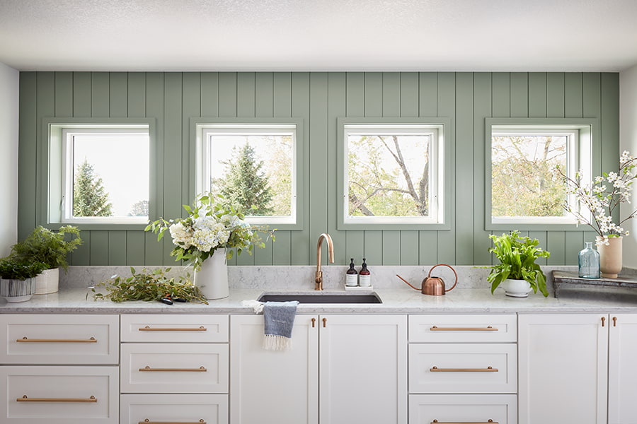Four awning windows on a green shiplap wall over a white kitchen counter and sink.