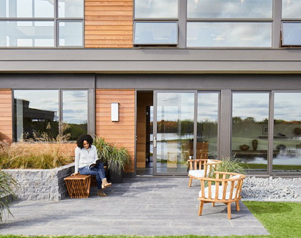 A woman is sitting on an outdoor patio in front of a contemporary home with custom windows and doors.