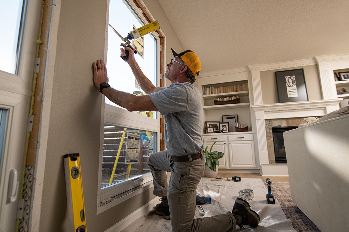 A man kneeling and installing a replacement window in a living room, demonstrating replacing a replacement window.