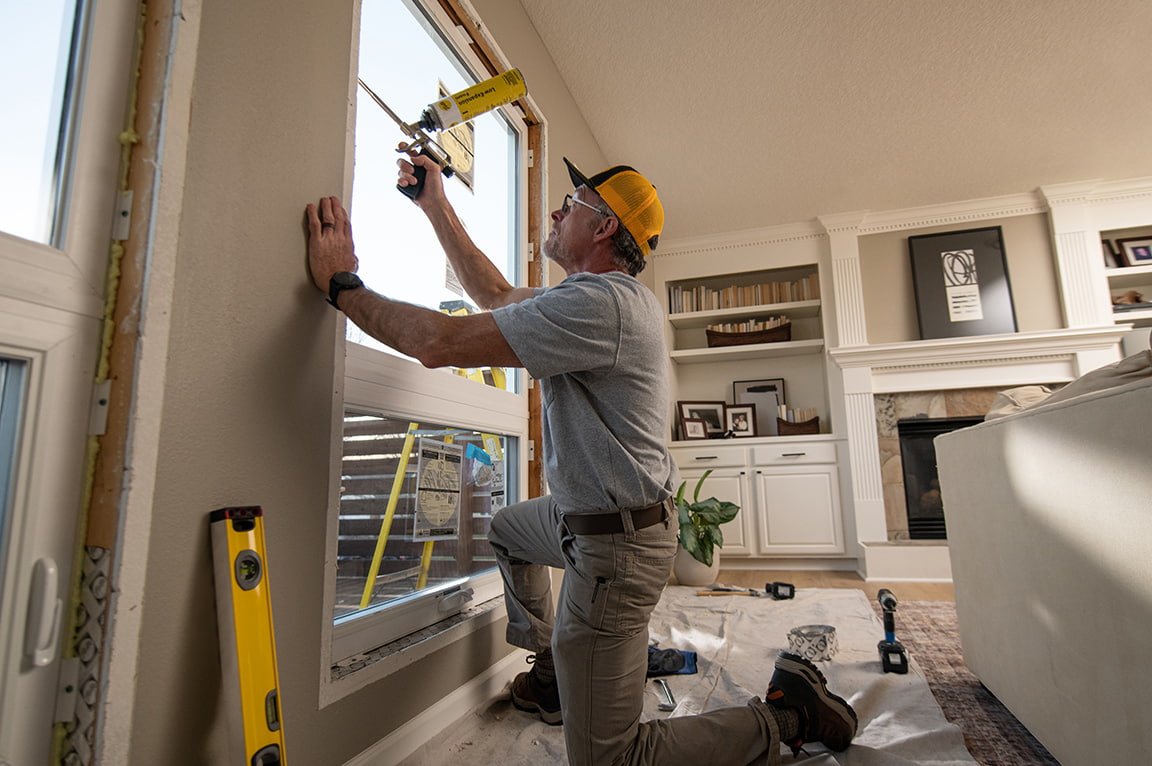 A man kneeling and installing a replacement window in a living room, demonstrating replacing a replacement window.