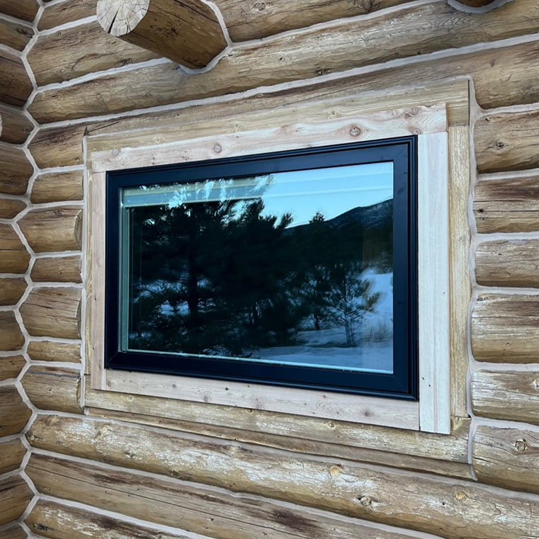 Black awning window in a wooden cabin, reflecting the sky and trees.