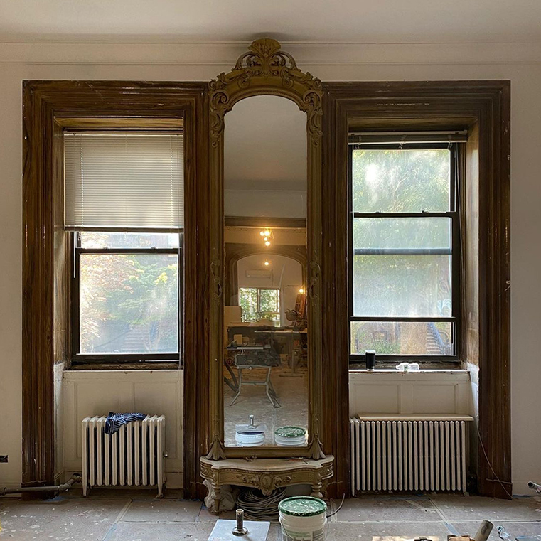 A before image of a room featuring brownstone windows and an ornate mirror frame, with renovation materials visible.