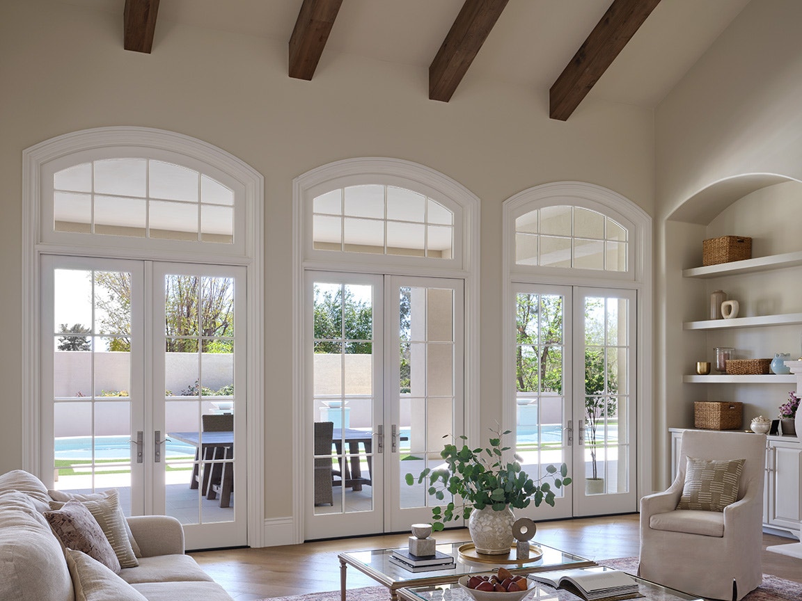A bright living room featuring arched transom windows, wooden beams, and a view of the outdoor patio.