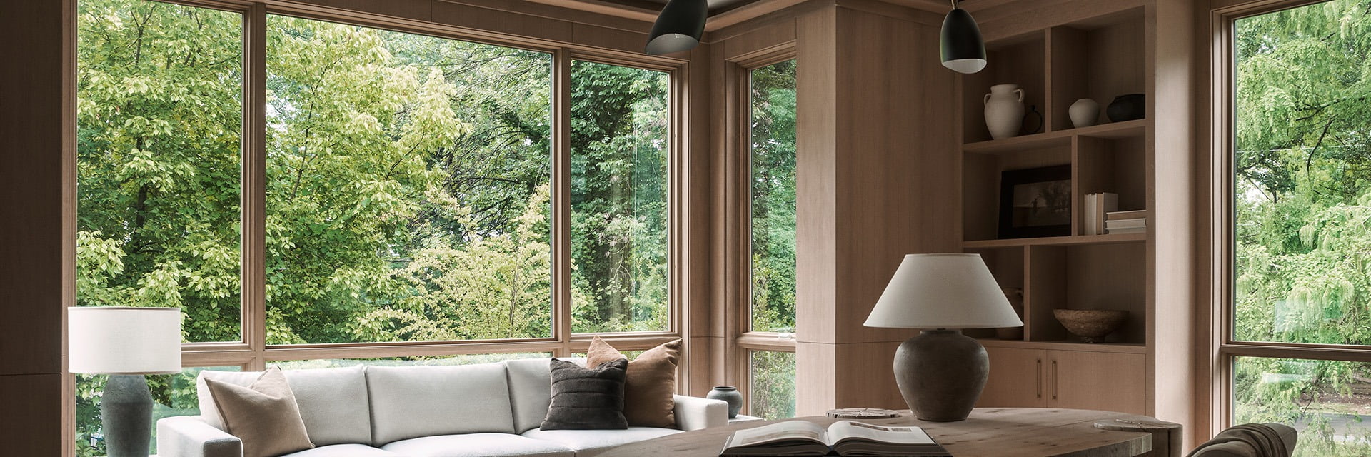 Warm home office featuring a wooden desk, neutral sofa, built-in shelves, and expansive wood-framed windows overlooking lush green trees.