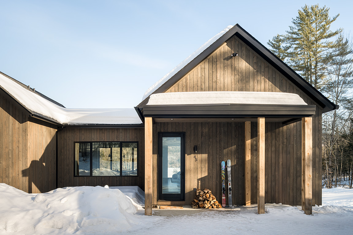 A modern cabin with a wooden exterior and fiberglass windows, surrounded by snow and trees.