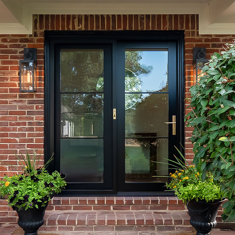 Modern black hinged entry door with full glass panel and brass handle on brick exterior.