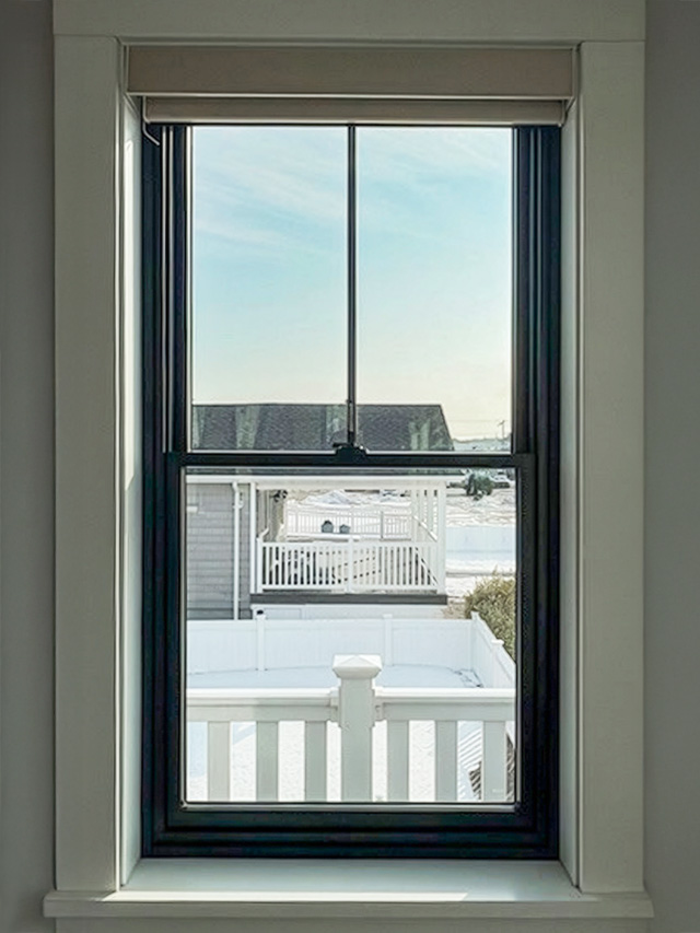 Black‑framed window in a bright interior space with a view of a fenced yard and water beyond.