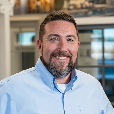 A man with brown hair and a brown beard is wearing a blue button-down shirt and smiling at the camera.