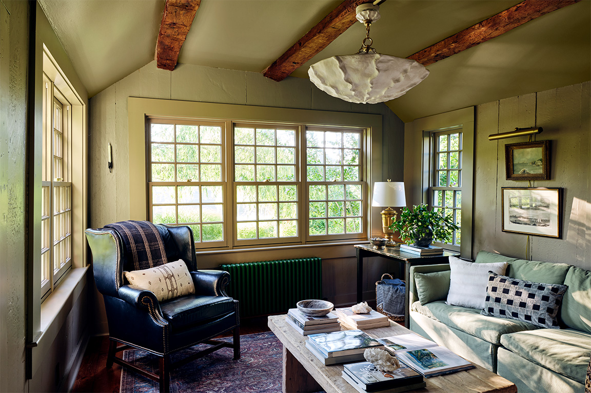 A cozy living room with large windows, featuring a blue armchair, wooden table, and decorative plants.