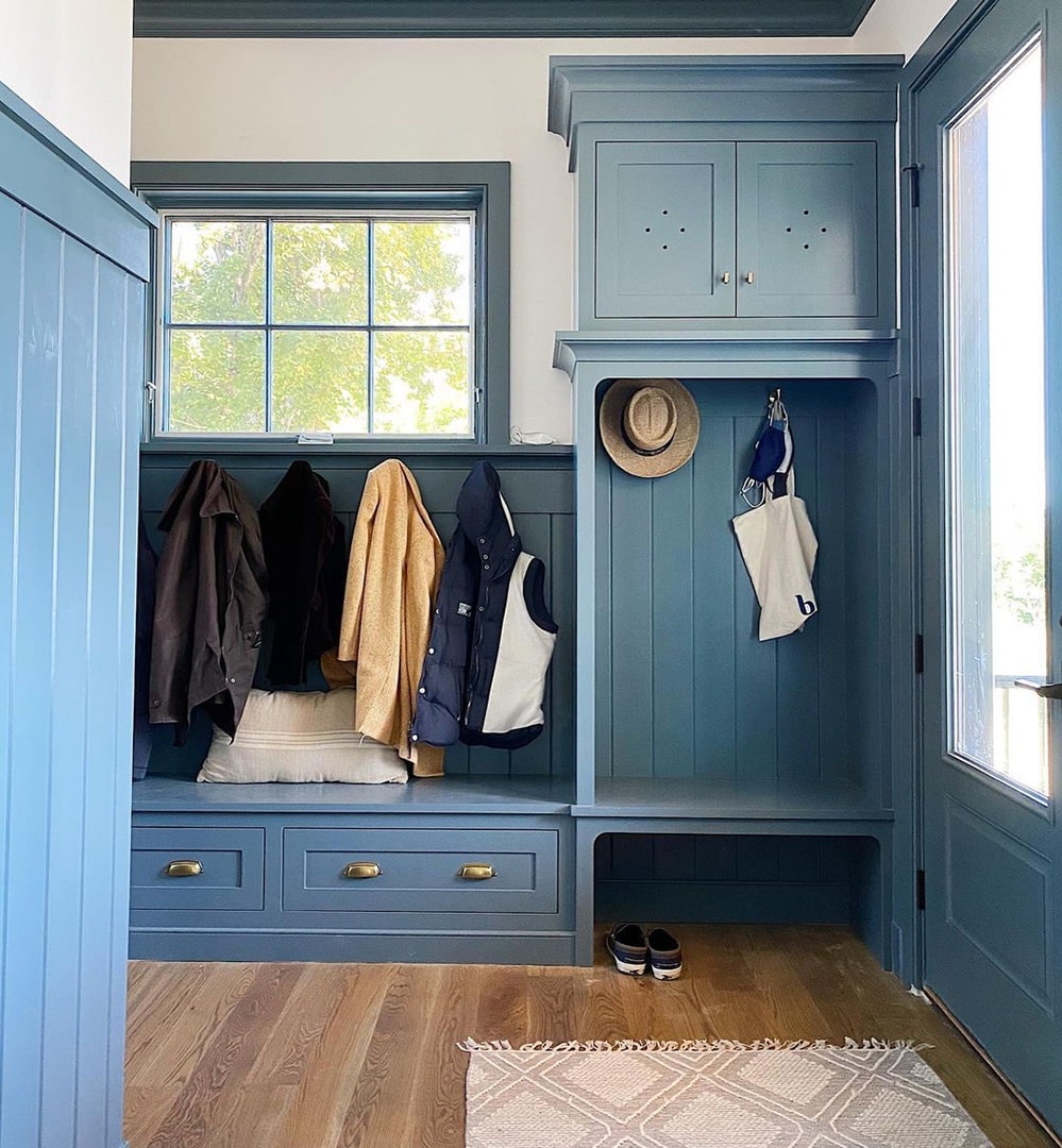 A mudroom with blue cabinets has an awning window above the coat hooks.