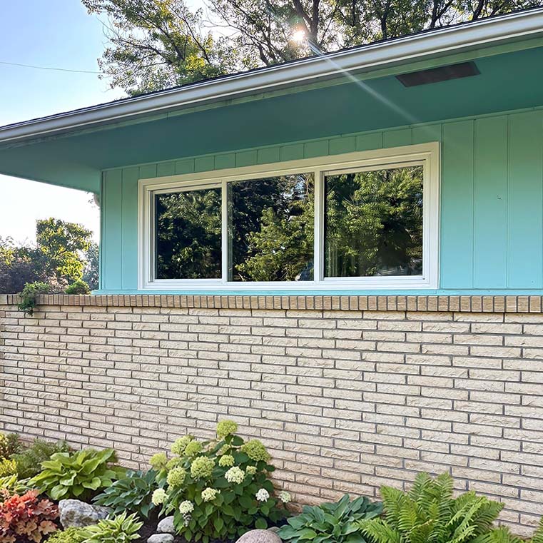 White-framed sliding window on a turquoise mid-century house above a tan brick wall, reflecting trees, with a landscaped hydrangea garden.