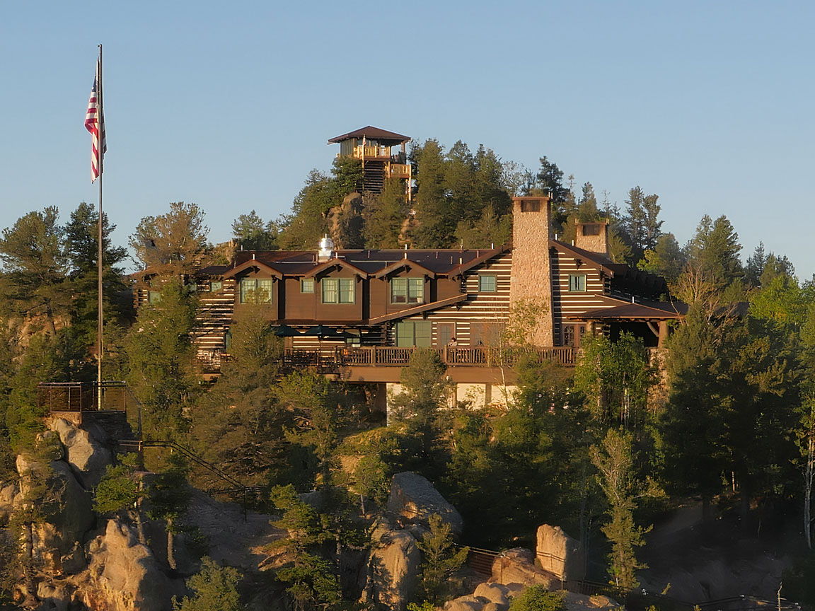 Large log cabin with multiple cabin windows and a stone chimney sits atop a forested mountain at sunset.
