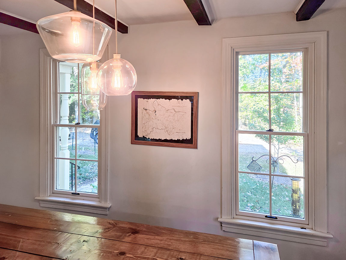 Dining room interior with two double-hung windows, white trim, and modern pendant lighting above a wooden table.
