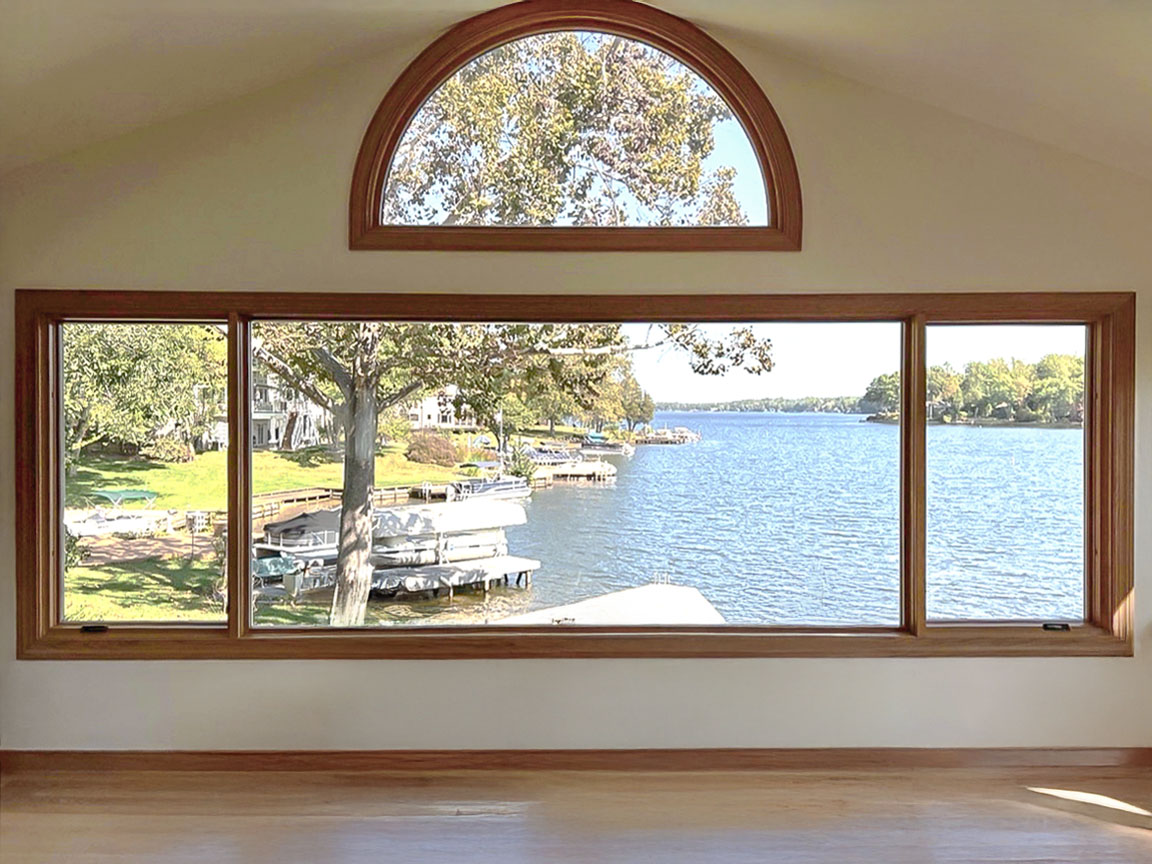 A large picture window with a semi-circular top section, offering a scenic view of a lake with boats docked along the shore and trees in the background.