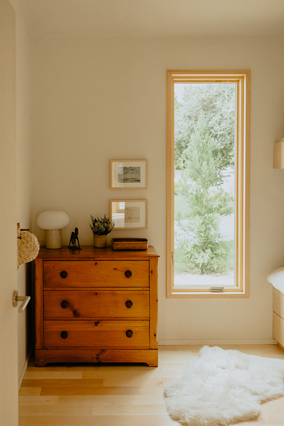 A tall energy efficient window with a wood frame brightens a cozy bedroom with a dresser and soft rug.