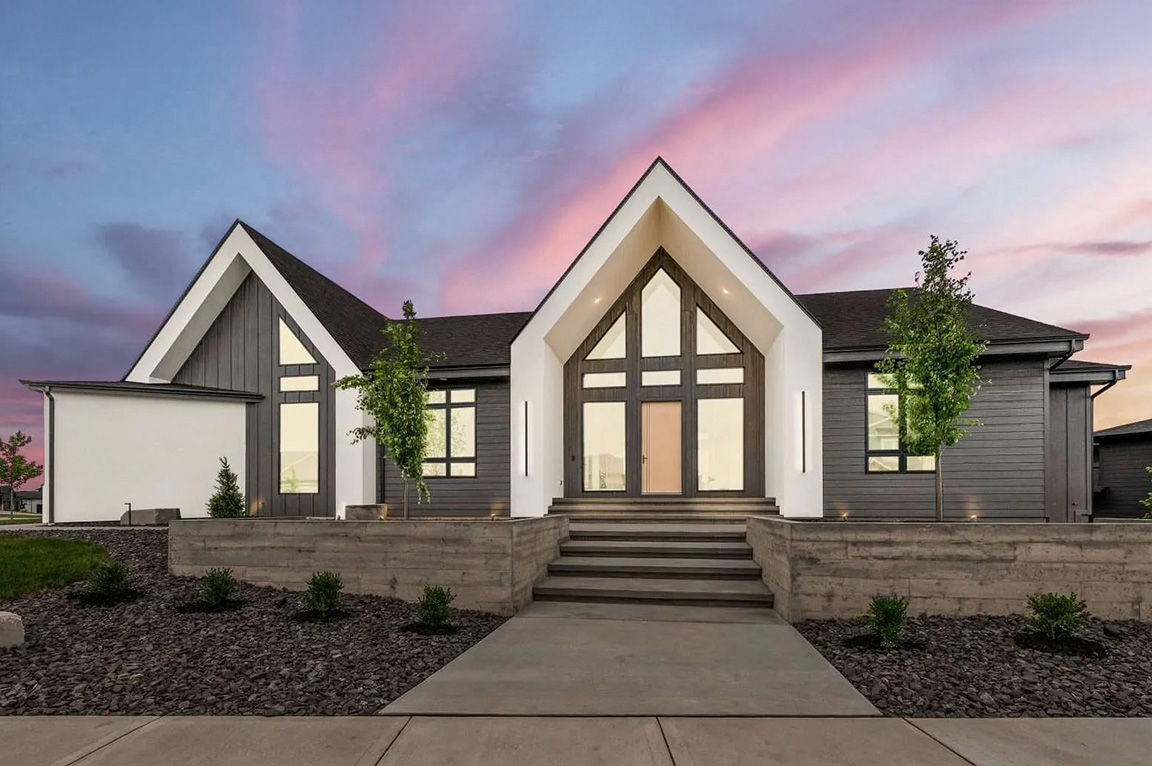 Modern gabled home exterior with gray siding, large windows, and dramatic sunset sky.