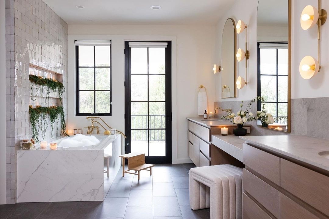 Between a marble bathtub and wood vanity are a black casement window and hinged patio door that leads to a deck.