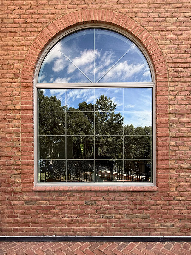 Arched window with tan brick surround reflecting trees and sky, showcasing Tan Window Replacement.