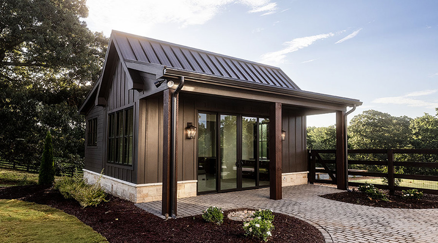 Modern dark-paneled pool house with metal roof, large glass sliding doors, and stone accents, surrounded by landscaped garden and a paved walkway