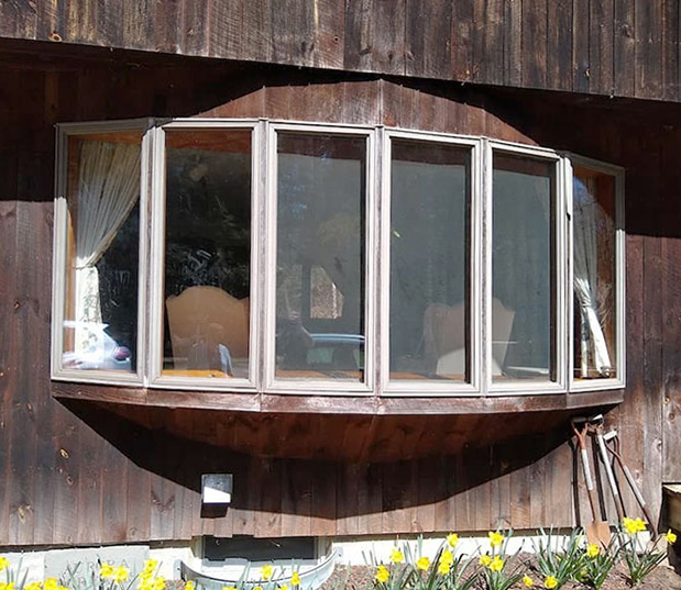 A bay window with red accents on a wooden house, surrounded by yellow flowers.