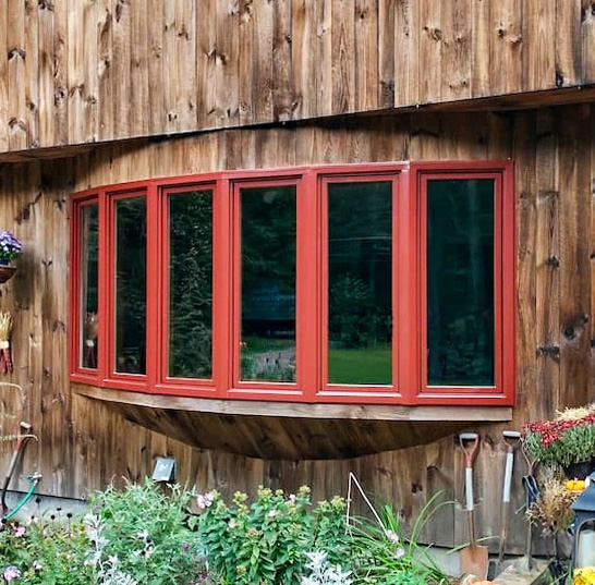 This rustic wood cabin features a bright red bow window that protrudes out from the wall.