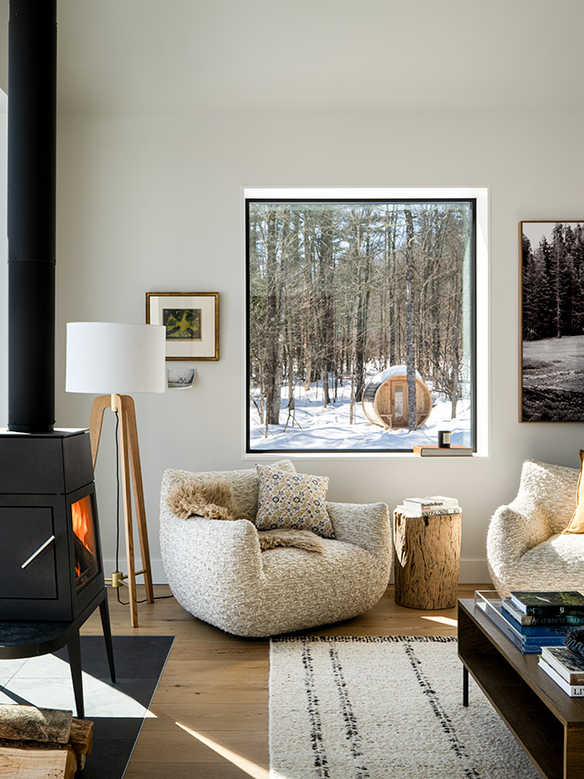 A cozy living room with fiberglass windows showcasing a snowy forest view, featuring a modern chair and a wood stove.