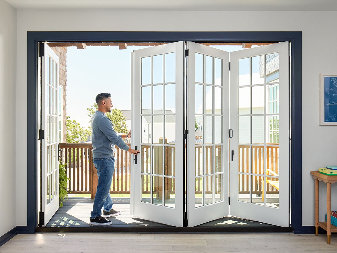 A man wearing a blue shirt is opening a large white bifold patio door.