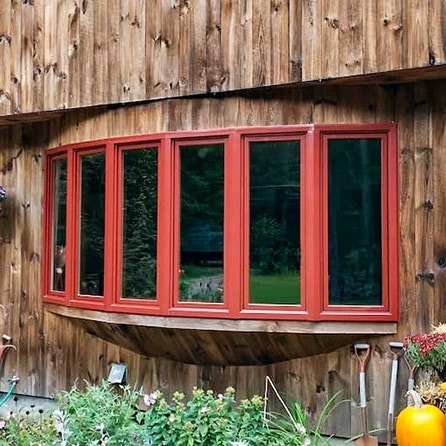 A red bow window replacement on a wooden house, surrounded by a garden with plants.