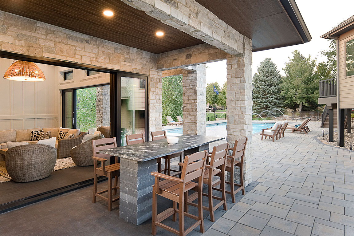 A modern pool house with large windows, featuring a stone patio and a dining space with wooden chairs.