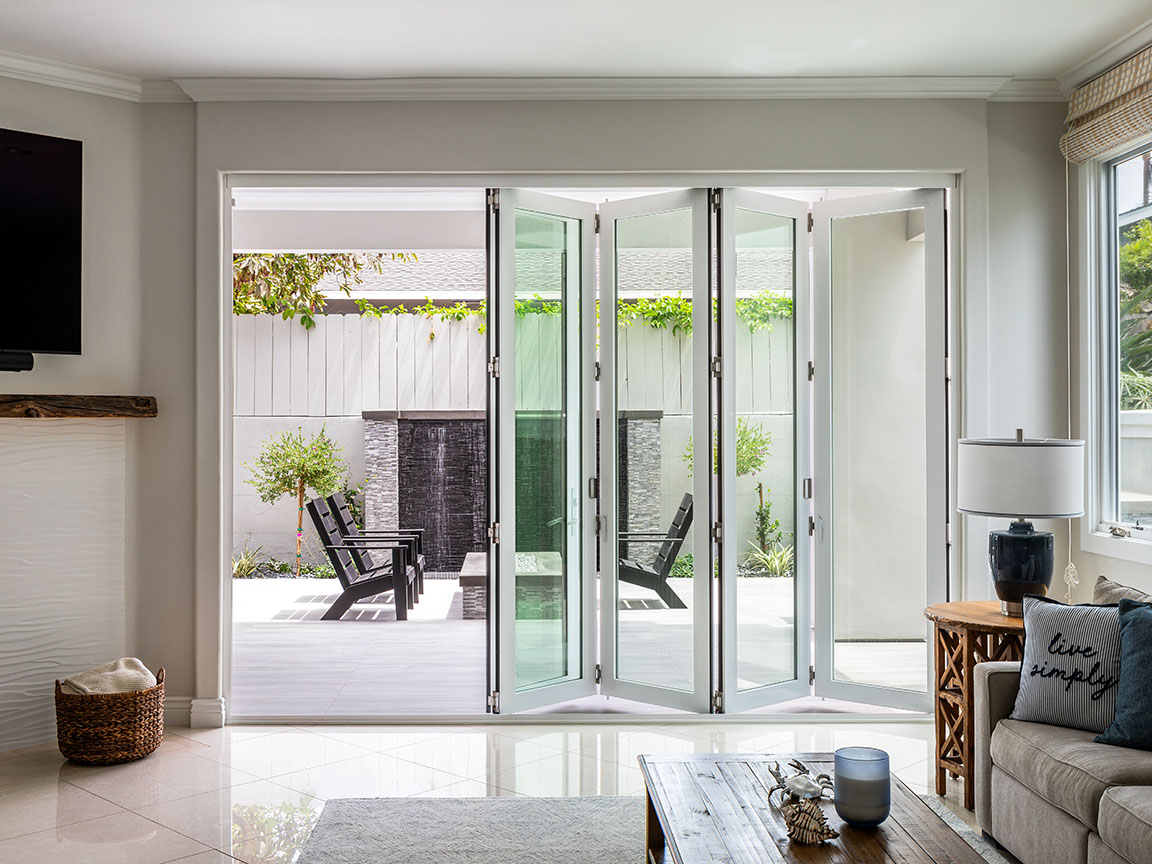 Large white bifold patio doors separate the minimalist living room from the exterior patio with greenery along the fence.
