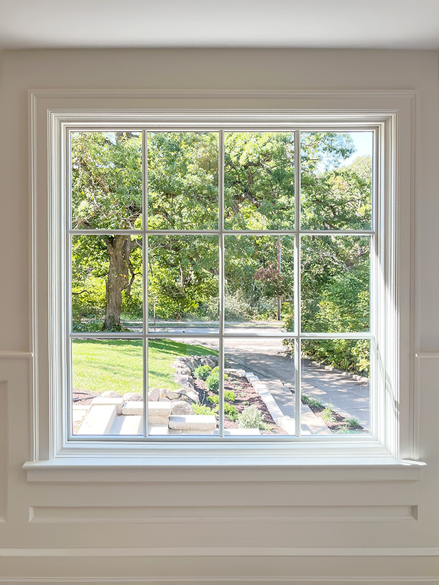 Large white-trim picture window showcasing a clear outdoor view of green trees, landscaped garden, and stone pathway on a sunny day.