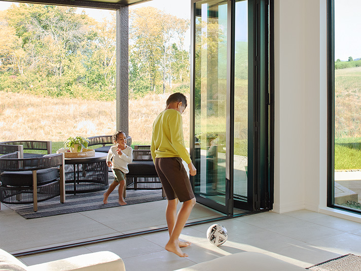 Children playing soccer indoors near an open bifold patio door leading to an outdoor seating area