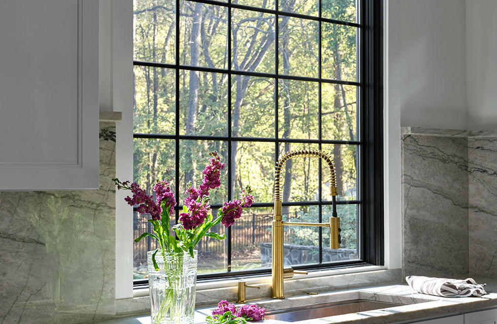 A large black fixed window with traditional grilles sits behind a farmhouse sink and a vase with purple flowers.