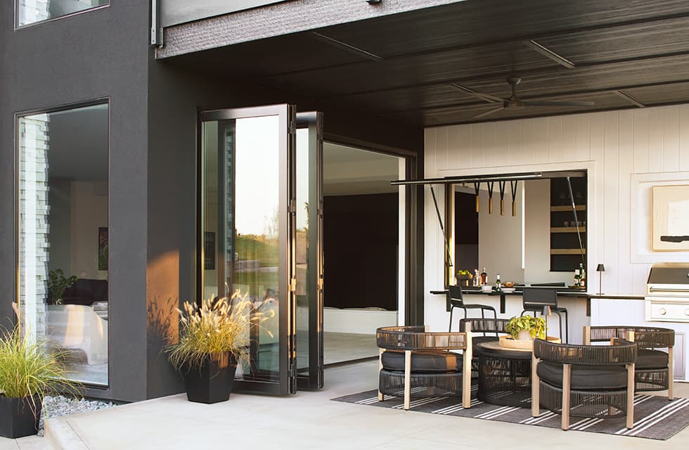 Contemporary patio with bifold doors, outdoor kitchen and grill, woven lounge chairs around round coffee table and potted grasses.