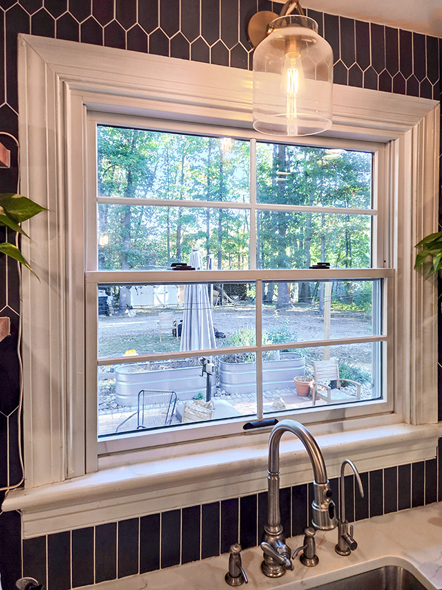 Kitchen interior featuring a double-hung window above a stainless steel sink, surrounded by black tile backsplash and modern light fixture.