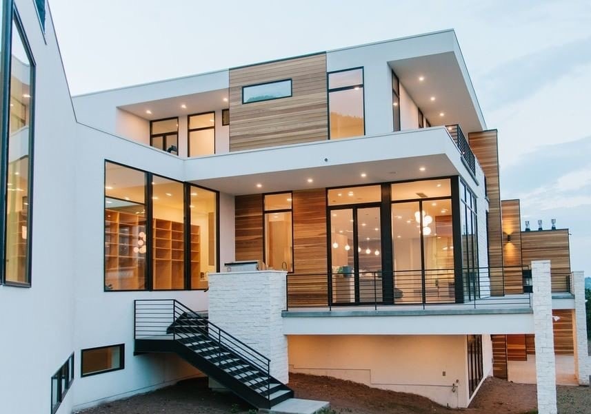 The exterior of a modern white home features black floor-to-ceiling windows and a large outdoor patio.