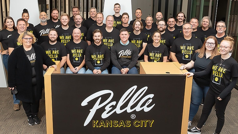 A large group of people in Pella shirts pose in a Kansas City windows showroom behind a branded sign.