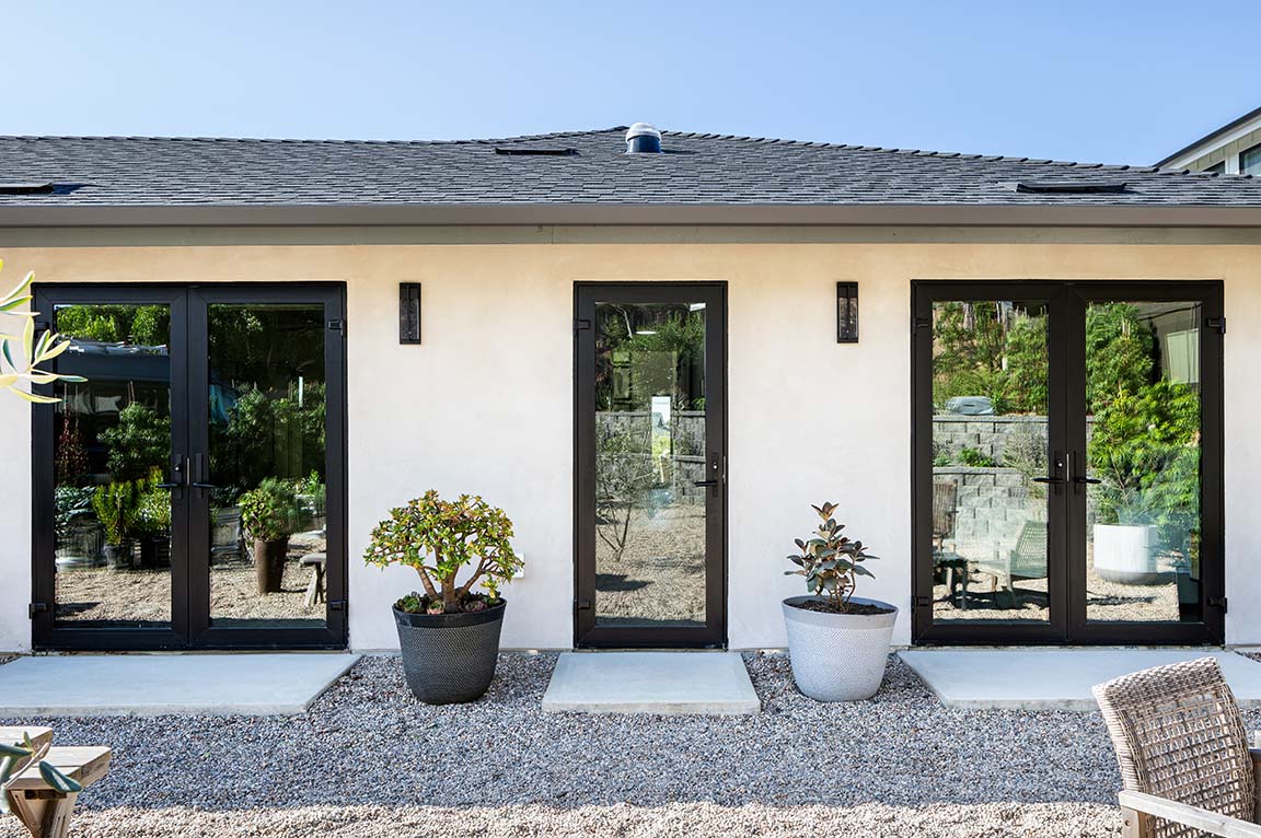 Three sets of black hinged doors on the back of a white home that open to a stone-filled back entertainment area.