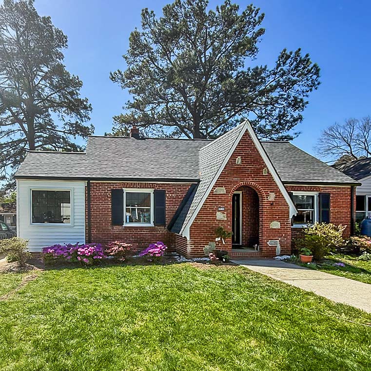 A charming brick cottage with a steeply pitched roof, arched entryway, and black shutters. The house is surrounded by a lush green lawn and vibrant flower beds, with tall trees in the background under a clear blue sky