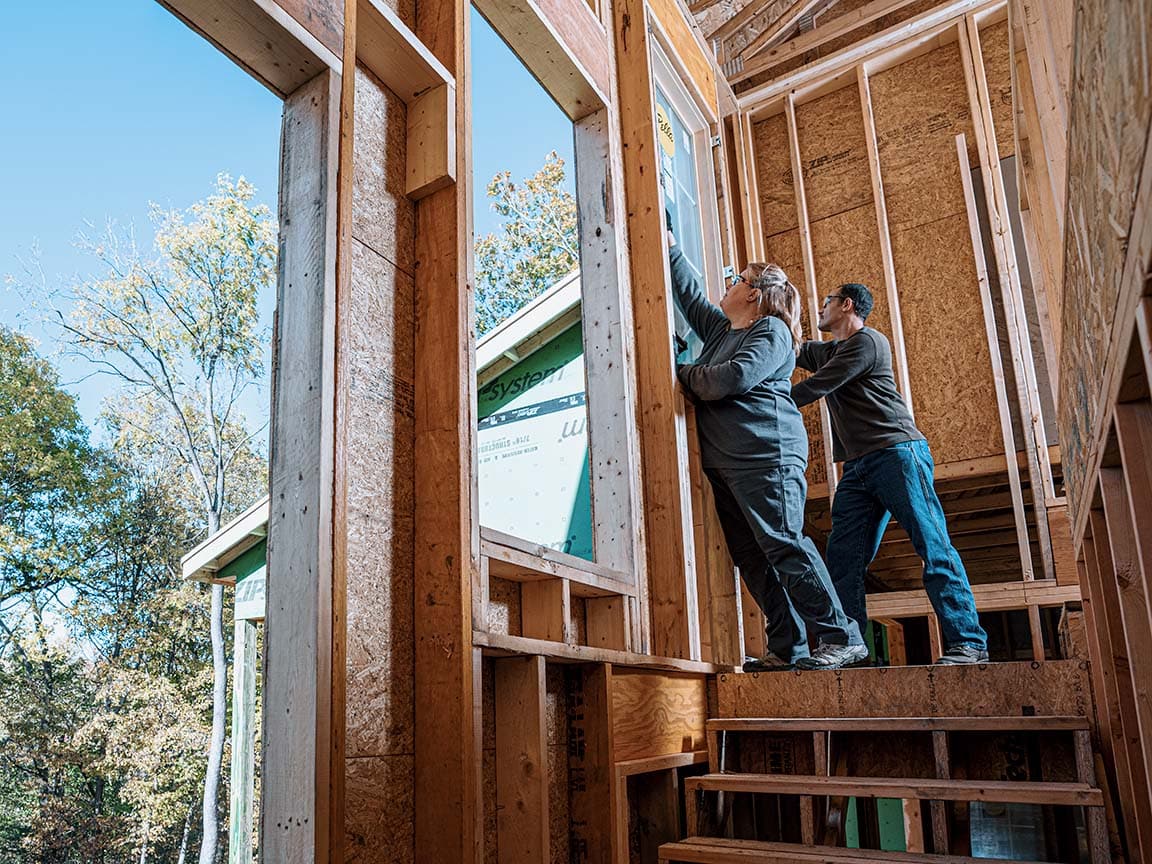 Two contractors are installing a new Pella window into a wood-framed home exterior.